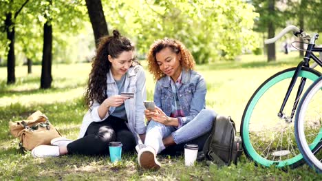 Two-young-women-are-paying-with-card-using-smartphone-sitting-in-park-on-grass-holding-credit-card-and-phone-then-doing-high-five-and-laughing.-Online-payment-concept.