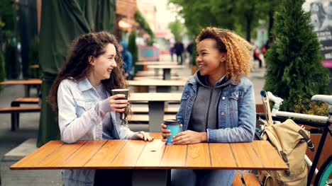 Zwei-junge-Frauen-trinken-Kaffee-und-reden,-sitzen-am-Tisch-im-Café-im-Freien-im-Park,-junge-Frauen-sind-Spaß-und-lachen.-Unterhaltung-und-Getränke-Konzept.