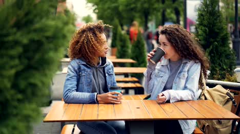 Fröhliche-Junge-Frauen-Beste-Freunde-lachen-und-reden-beim-Sitzen-am-Tisch-im-Café-im-freien-und-Kaffee-zu-trinken,-sind-Mädchen-Geselligkeit-und-Spaß-Neuigkeiten-diskutieren.