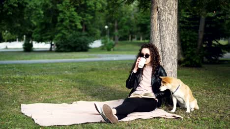 Relaxed-girl-is-reading-book-and-drinking-takeaway-coffee-sitting-on-lawn-in-park-while-her-cute-dog-is-smelling-ground-and-air-sitting-under-tree.-Leisure-and-pets-concept.