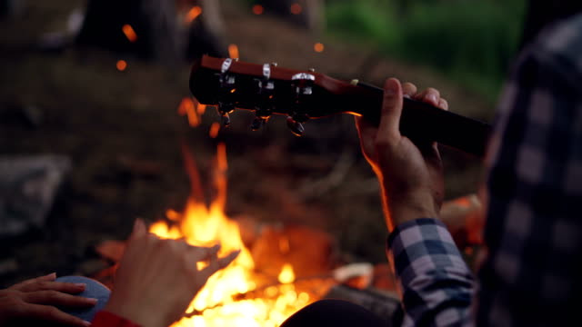 Close-up-shot-of-male-hands-playing-the-guitar-and-female-hands-moving-in-dance-with-burning-campfire-in-background.-Camping,-music-and-people-concept.
