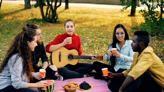 Grupo-multiétnico-de-alegres-jóvenes-son-tintineantes-vasos-con-bebidas-y-beber-sentado-en-manta-en-el-césped-durante-otoño-picnic-con-comida-y-guitarra.