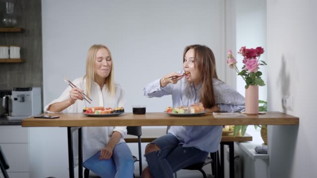 Two-young-women-talking-and-eating-sushi-at-home,-best-friends-spending-time-together