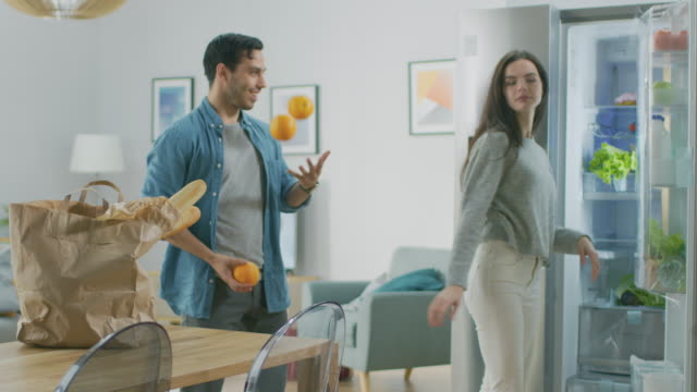 Beautiful-Young-Couple-Have-Fun-in-the-Kitchen.-Girl-Puts-Fresh-Salad-Greens-Inside-the-Modern-Fridge.-Man-is-Juggling-with-Oranges.-She-Cheers-for-Him-and-They-Laugh.