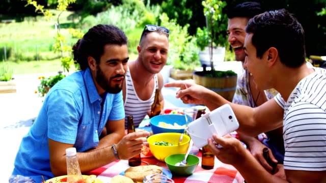 Lächelnder-Mann-zeigt-sein-Handy-zu-seinen-Freunden-beim-Essen-im-Freien-mit