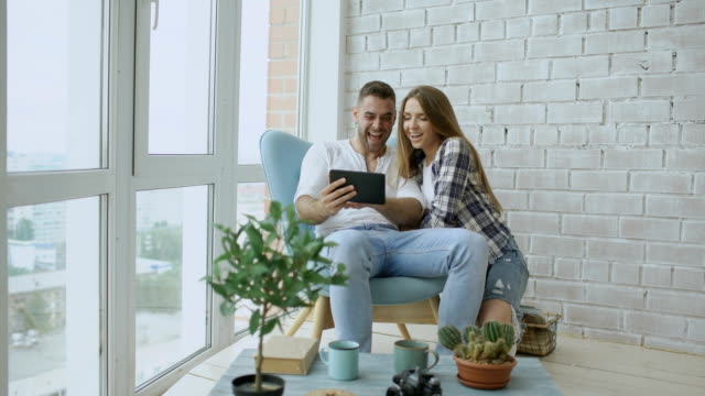 Young-attractive-couple-having-online-video-chat-using-tablet-computer-sitting-at-balcony-in-modern-loft-apartment
