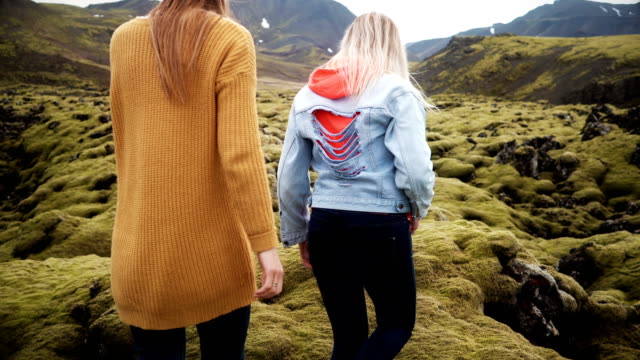 Back-view-of-two-tourists-woman-hiking-together-in-Iceland.-Girls-walking-through-the-lava-field-covered-moss