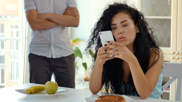 Woman-uses-phone-sitting-at-table,-funny-man-squats-at-the-background
