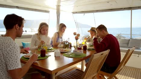 Gruppe-von-jungen-schönen-Menschen-essen-gesundes-Frühstück-auf-einer-Yacht,-sie-sind-freundlich-im-Chat.-Sie-haben-verschiedene-Gerichte-und-Smoothies-auf-den-Tisch-serviert.-Meer-ist-im-Hintergrund.