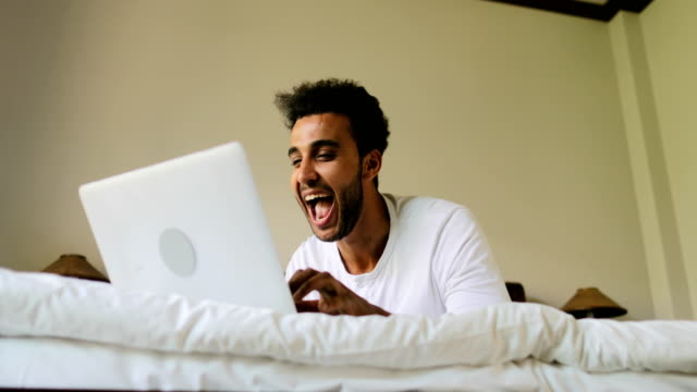 Junger-Mann-mit-Laptop-Computer-auf-Bett-glücklich-lächelnd-Hispanic-Kerl-im-Chat-Online,-Schlafzimmer-morgens