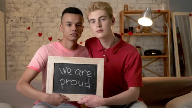 A-sad-international-gay-couple-is-sitting-on-the-couch-and-holding-a-sign.-We-are-proud.-Look-at-the-camera.-Home-comfort-on-the-background.-60-fps