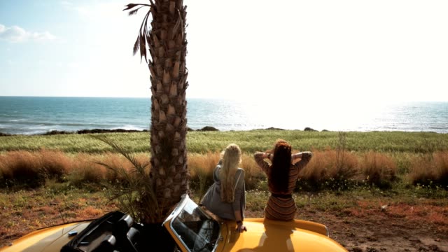 Young-women-sitting-on-retro-convertible-car-looking-at-sea