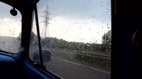 View-through-the-side-window-of-the-old-retro-car-while-moving-at-rainy-weather.-Raindrops-falling-on-the-glass-of-vintage-automobile-during-drive-at-countryside.-Close-up-Slow-motion