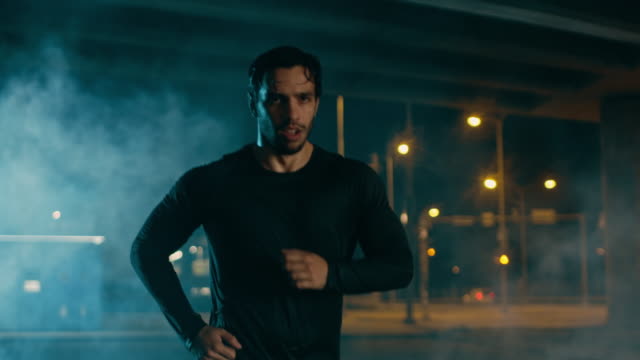 Sweating-Athletic-Muscular-Young-Man-in-Sports-Outfit-Jogging-in-a-Street-Filled-With-Smoke.-He-is-Running-in-an-Evening-Urban-Environment-Under-a-Brindge-with-Cars-in-the-Background.