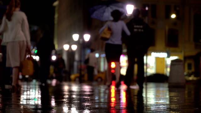 Abstract-background-rainy-late-evening-in-the-city-in-naturale-dark-tones.-Raindrops-fall-on-the-colorful-asphalt-illuminated-by-street-lamps.-Among-the-crowd-of-pedestrians-go-two-friends-under-umbrellas-and-talk.-Lifestyle-of-modern-city