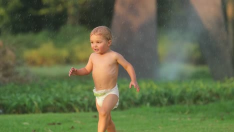 Beautiful-real-life-moment-of-infant-baby-boy-running-in-slow-motion-60fps-in-green-field-with-water-sprinkes-on-during-summer-sunset-golden-hour-time-in-4k-clip-resolution