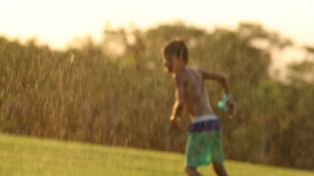 Candid-Traumszene-der-Kind-spielt-mit-Wasser-besprengt-bei-Sonnenuntergang.-Junge-springen-und-Spaß-mit-den-einfachen-Dingen-im-Leben,-ein-Wasser-streuen-in-den-Rasen-in-4k-Auflösung-Clip