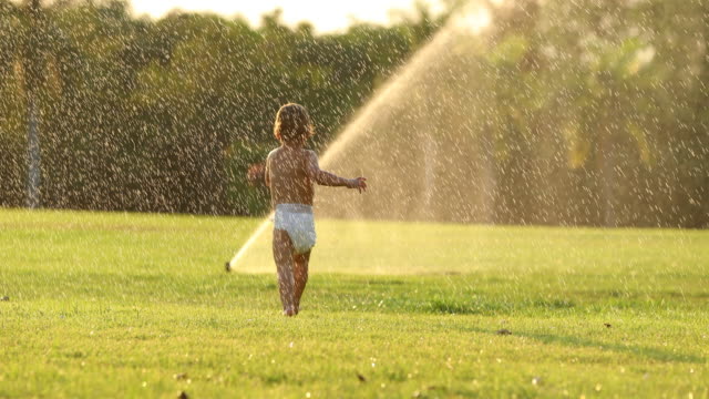Dreamy-idealic-scene-of-infant-toddler-boy-running-in-green-field-with-water-sprinkles-during-sunset-golden-hour-time-in-4k-clip-resolution
