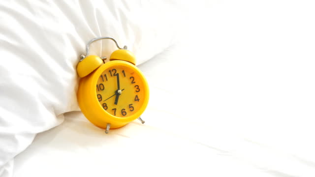 Yellow-alarm-clock-ringing-on-white-bed.-Holiday-and-Vacation-concept.-Object-and-Home-theme.-Close-up-panning-to-clock.-pillow-and-blanket-elements.-4k-Living-crop