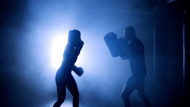 Silhouettes-of-a-woman-boxer-trains-her-punches-on-a-punching-bag-that-her-partner-in-dark-gym