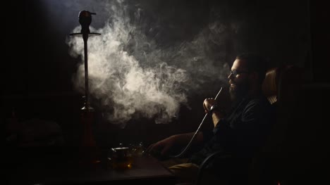 young-bearded-man-in-glasses-smokes-a-hookah-and-blow-out-smoke-closeup-on-black-background-in-slow-motion-in-4k