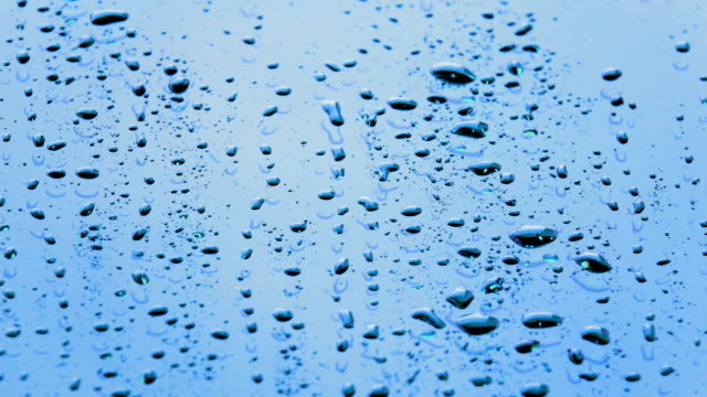 Rain-droplets-or-water-droplets-flowing-on-glass-on-sunlight-daytime.-Closeup-rain-falling-to-splash-on-bright-surface-of-mirror-of-car-background
