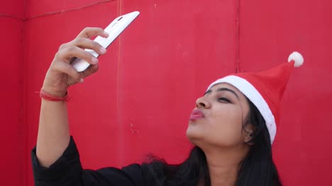 Indische-Frau-mit-Nikolausmütze-Selfies-Foto-mit-Handy,-fröhlich-und-aufgeregt,-mit-rotem-Hintergrund