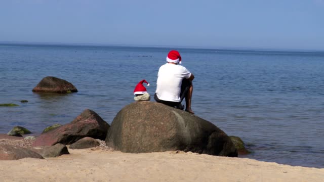 Mann-sitzt-auf-Felsen-am-Meer-mit-Weihnachtsmann-Mütze