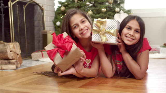 Two-sisters-lay-on-the-floor-in-front-of-the-christmas-tree-and-shake-their-presents.-Portrait