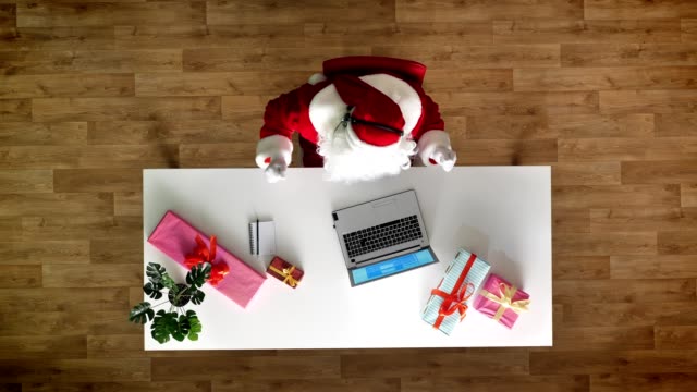 Santa-Claus-working-at-office-on-laptop,-listening-music-in-headphones,-typing,-aerial-view,-top-down-shot,-writes-text-to-a-notebook