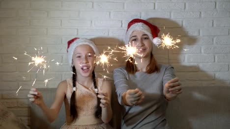 Two-sisters,-a-little-girl-and-a-teenager,-in-Santa's-hats-holding-lighted-sparklers-in-their-hands,-wave-them-and-smile-sitting-on-the-couch-on-a-white-background.