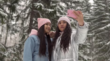 Two-young-girlfriends-in-winter-clothes-taking-selfie-against-the-background-of-the-winter-forest-using-a-smartphone-and-smiling.-4K