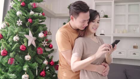 Asian-couple-using-smartphone-selfie-with-christmas-tree-decorate-their-living-room-at-home-in-Christmas-Festival.-Lifestyle-woman-and-man-happy-celebrate-summer-christmas-and-New-year-concept.