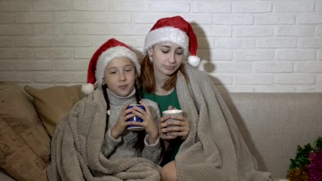 Zwei-kleine-Mädchen-in-Santas-Hüte-sitzen-mit-einer-Decke-bedeckt-und-halten-die-Becher-in-der-Hand-gegen-eine-weiße-Mauer-auf-der-Couch.-Sie-tanzen-und-Lächeln.