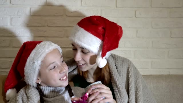 Retrato-de-dos-niñas-en-Santa-Claus-sombreros-cubiertos-con-una-manta,-se-sientan-en-el-sofá,-sosteniendo-tazas-y-sonriendo.-De-cerca.-4-K.-25-fps.