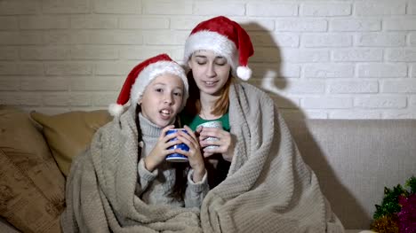 Kinder,-zwei-Mädchen,-mit-einer-Decke-bedeckt-singen-im-Weihnachtsmann-Mützen-auf-der-Couch-gegen-eine-weiße-Wand-im-flackernden-Licht-sitzen.-Porträt.-4-K.-25-fps.