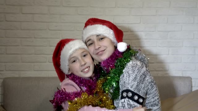 Portrait-of-two-happy-children-girls-in-Santa-Claus-hats-with-Christmas-tinsel-on-their-shoulders,-they-sit-on-the-couch,-hug,-look-at-the-camera-and-smile.-Close-up.