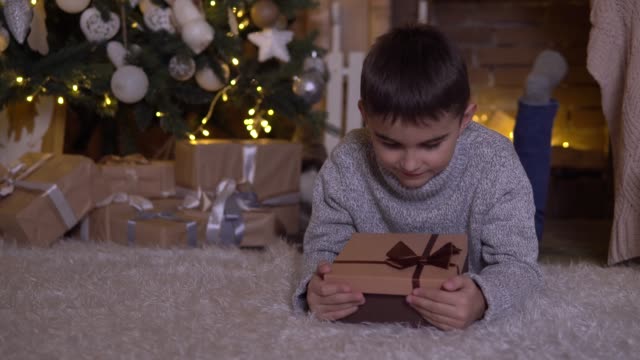 A-little-boy-opens-a-box-with-a-gift-and-rejoices-lying-on-the-floor-near-the-Christmas-tree.-4K