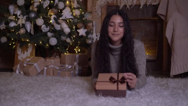 La-chica-abre-la-caja-con-un-regalo-y-regocija-en-el-suelo-cerca-del-árbol-de-Navidad