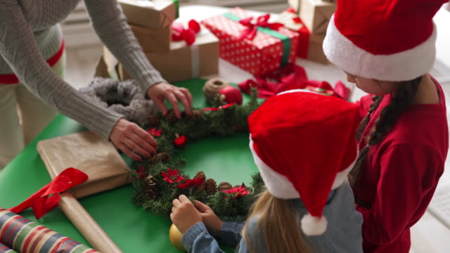 Zwei-Mädchen-in-Santa-Hüte-und-Frau-machen-Weihnachten-Kranz