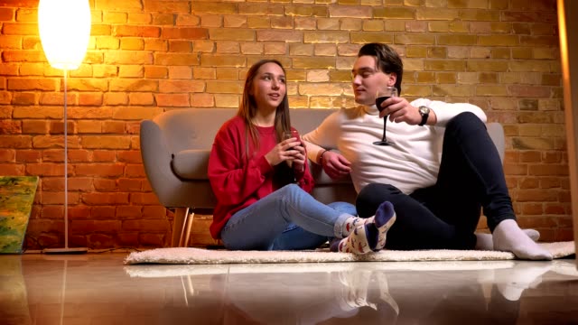 Portrait-of-young-caucasian-friends-sitting-on-carpet-and-drinking-wine-talking-with-each-other-in-cosy-home-atmosphere.