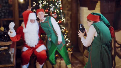 Ein-Assistent-Elf-ist-Santa-Claus-und-eine-Elfe-auf-dem-Hintergrund-von-einem-Weihnachtsbaum-und-Girlanden-am-Fenster-mit-einem-Retro-Kamera-auf-Vorabend-des-neuen-Jahres-fotografieren.