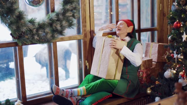 Cansado-elfo-con-orejas-largas-está-sentado-en-el-alféizar-de-la-ventana-con-un-regalo-en-la-mano-en-el-fondo-de-la-ventana-cerca-del-árbol-de-Navidad-antes-del-año-nuevo.