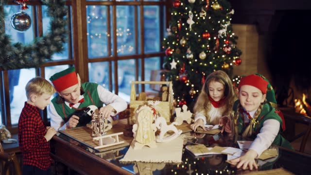 Two-elves-are-sitting-at-the-table-with-a-little-boy-and-girl-and-show-them-letters-of-congratulations-and-Christmas-gifts-on-the-background-of-the-Christmas-tree-and-the-fireplace-near-the-window-in-the-residence-of-Santa-Claus