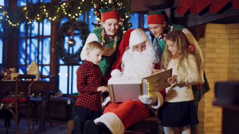 Santa-Claus-está-sentado-en-una-silla-rodeada-por-dos-duendes-y-un-niño-pequeño-con-una-chica-y-teniendo-en-cuenta-un-álbum-de-vacaciones-con-fotos-en-el-fondo-de-adornos-de-año-nuevo-y-el-árbol-de-Navidad-en-la-sala-en-casa