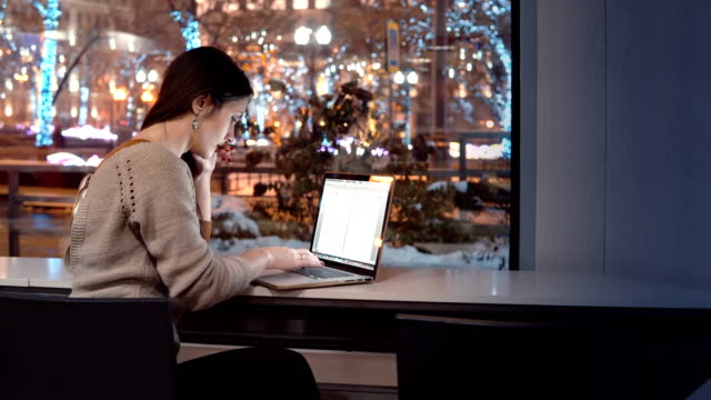 Atractivo-joven-empresaria-trabajando-en-ordenador-portátil-sentado-en-el-bar,-fuera-de-ciudad-de-la-noche-de-invierno-decorado-para-la-Navidad
