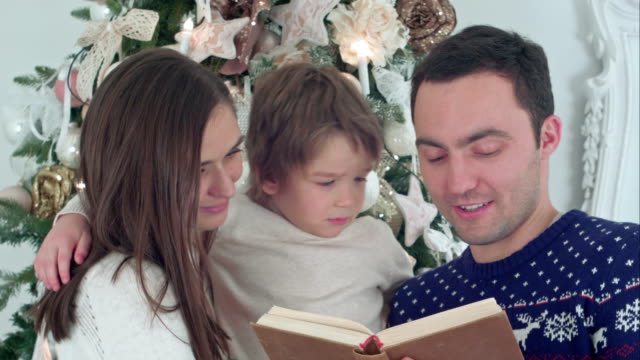 Joven-padre-leyendo-cuento-de-Navidad,-mientras-que-la-madre-y-su-hijo-feliz-susurrando-secretamente