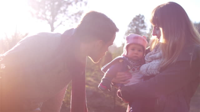 A-young-dad-talks-to-his-baby-girl-while-mom-holds-her,-at-a-Christmas-tree-farm,-lens-flare