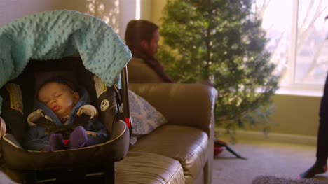 El-bebé-duerme-en-su-asiento-de-coche-en-el-sofá-mientras-que-mamá-y-papá-establecen-el-árbol-de-Navidad