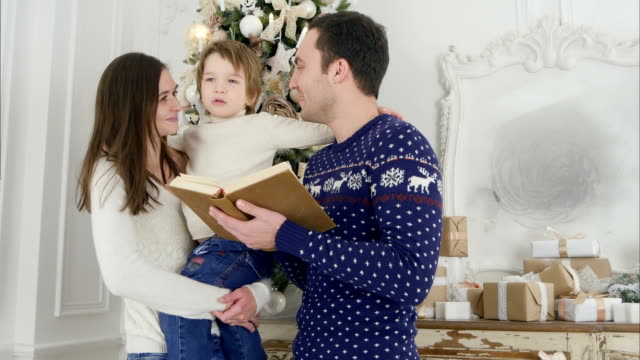 Glücklicher-Vater-liest-ein-Weihnachtsmärchen,-während-die-Mutter-ihren-fröhlichen-Sohn-vor-dem-Weihnachtsbaum-hält
